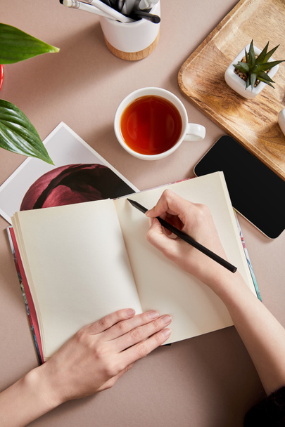 καλλιεργημένη άποψη της γυναίκας που γράφει στο σχεδιασμό κοντά σε πράσινα φυτά σε ξύλινο πίνακα, φλιτζάνι τσάι, smartphone σε μπεζ επιφάνεια - Φωτογραφία, εικόνα