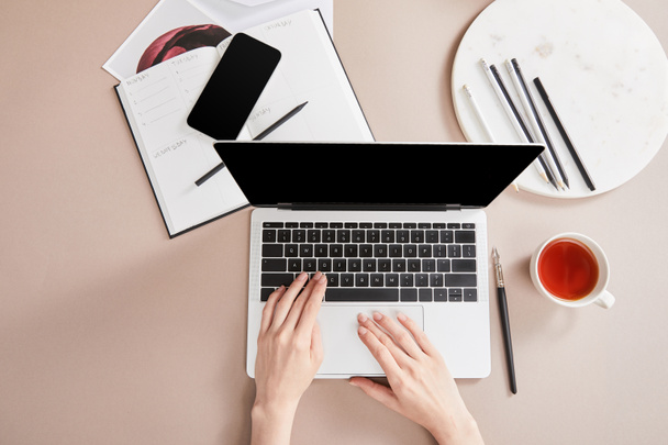 rajattu näkymä nainen käyttää kannettavaa tietokonetta lähellä kuppi teetä, suunnittelija, älypuhelin lähellä ympyrää kyniä beige pinnalla - Valokuva, kuva