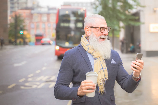 Empresário sénior feliz a beber café e a sair do trabalho. Homem em um dia chuvoso no meio do outono. Homem esperando o ônibus e olhando smartphone. Conceito de estilo de vida de trabalho e moda - Imagem
 - Foto, Imagem