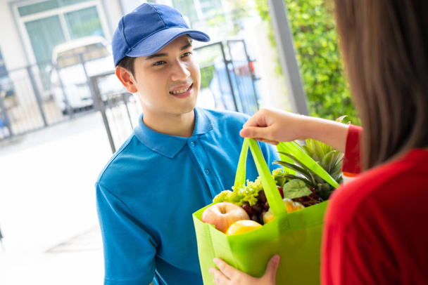 Food Deliver asiatische Mann in blauen Uniform geben Obst und Gemüse an Empfänger Kundenfront, Fast Express-Lebensmittelservice, wenn Krise Coronavirus, covid19 neue normale Lebensstil-Konzept. - Foto, Bild