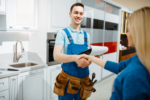 Male plumber in uniform and female customer in the kitchen. Handyman with toolbag repair sink, sanitary equipment service at home - Фото, зображення