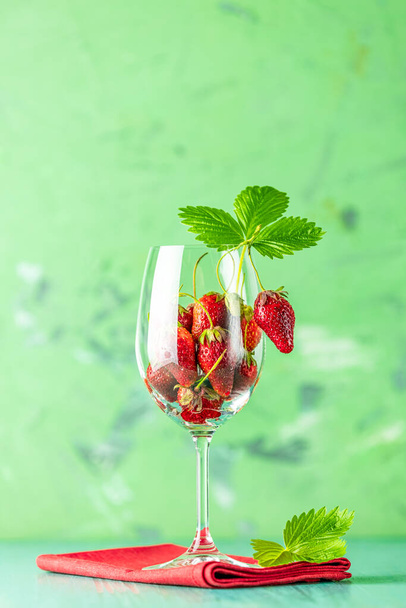 Fresh ripe raw strawberry with green leaves in the wine glass on green surface with red napkin. - Foto, imagen