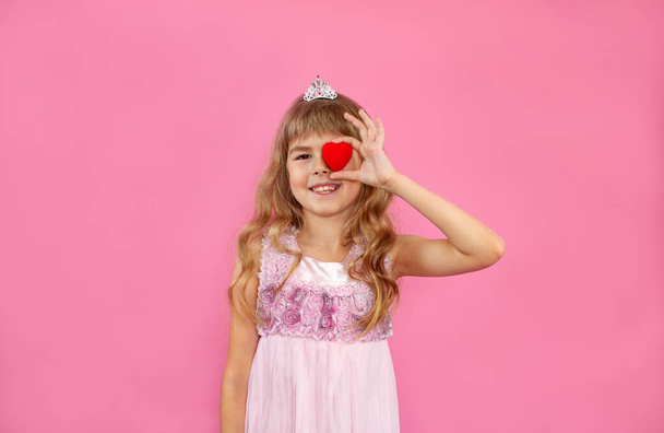 Little girl on a pink background. Little girl with a heart in her hands. Little girl is looking with love. The child holds a heart.Childhood happiness. Happy little girl.Girl in a beautiful dress. - Photo, Image