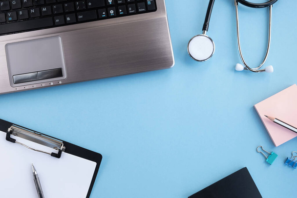 Ärztlicher Arbeitsplatz. Medizinisches Stethoskop, Laptop, leeres Klemmbrett und Stift auf hellblauem Hintergrund. Draufsicht, flache Lage, Kopierraum - Foto, Bild