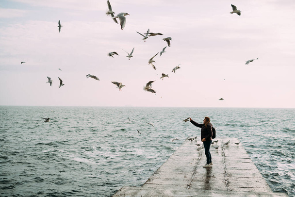  A girl stands near the sea on a pier with a backpack and feeds the seagulls. - Zdjęcie, obraz