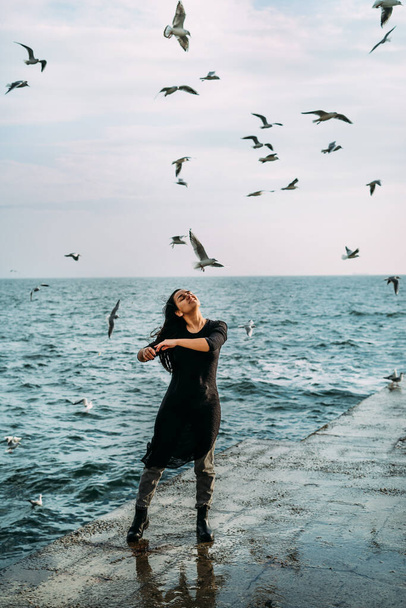 The girl in black stands on the pier with open arms to the sun and the wind receives inspiration and strength and inner peace of the soul seagulls. Open to the challenges and opportunities of life - Foto, afbeelding