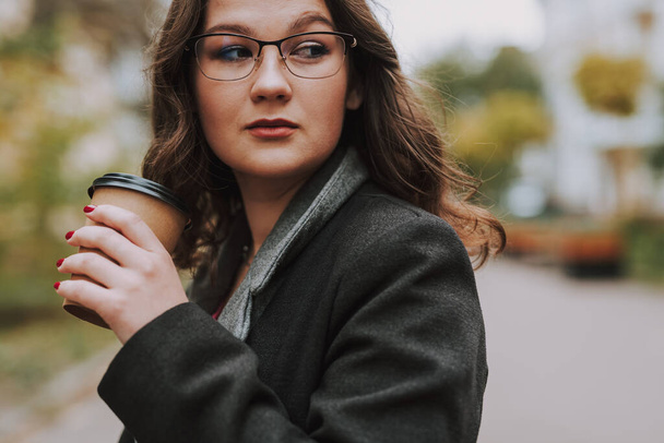 Muotokuva nuoresta naisesta takissa, joka pitelee kahvikuppia ja harkitsee kaukaisuutta. - Valokuva, kuva