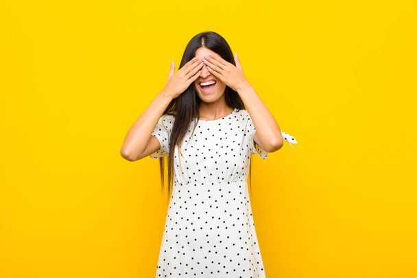 young pretty latin woman smiling and feeling happy, covering eyes with both hands and waiting for unbelievable surprise against flat wall - Photo, image