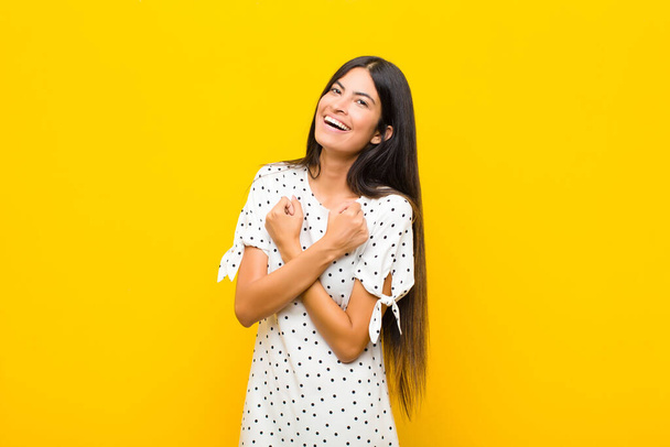 young pretty latin woman smiling cheerfully and celebrating, with fists clenched and arms crossed, feeling happy and positive against flat wall - 写真・画像