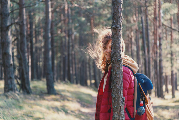 Young pretty woman walks in the forest, smiles and hides behind a pine trunk, local travel trend - Фото, изображение