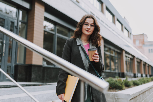Ruhige lächelnde Frau mit einer Papiertasse Kaffee und einem Buch in der Hand, während sie draußen auf der Treppe eines modernen Bürozentrums steht - Foto, Bild