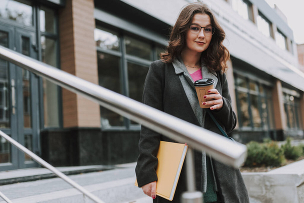 Kedves fiatal hölgy áll a szabadban, egy modern épület mellett, és mosolyog, miközben iszik egy csésze kávét. - Fotó, kép