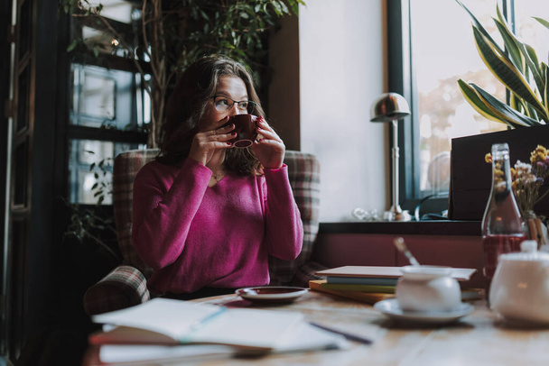 Pencerenin yanında oturan genç bir kadın işten sonra kafede dinlenirken elinde çayla fincanı tutuyor. - Fotoğraf, Görsel