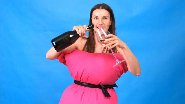 The most beautiful girl in a pink dress from pillows pours champagne into a glass on a blue background. Crazy quarantine. Fashion 2020. Put on a pillow. Challenge 2020 due to house isolation. - Фото, зображення