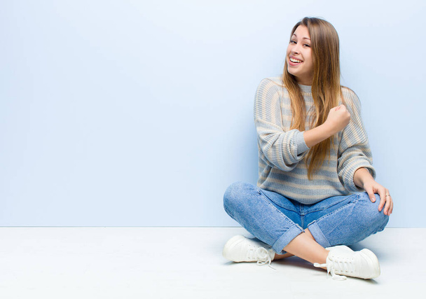 young blonde woman feeling happy, positive and successful, motivated when facing a challenge or celebrating good results sitting on the floor - 写真・画像