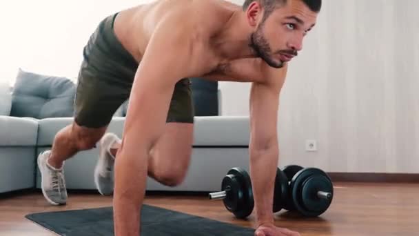 Genç adam evde egzersiz yapıyor. Kalıcı adam yoga minderinin üzerinde tahta pozisyonunda durur ve dağcı egzersizi yapmaya başlar. Bacakları hızlı ve tempoyla hareket ediyor. - Video, Çekim
