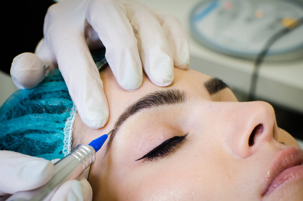 Kosmetikerin schminkt Augenbrauen permanent, junge Frau im Schönheitssalon tätowiert, extreme Nahaufnahme - Foto, Bild