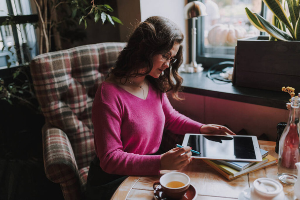 Lächelnde junge Frau mit Brille und Blick auf den digitalen Tablet-Bildschirm, während sie am Tisch im Café sitzt - Foto, Bild