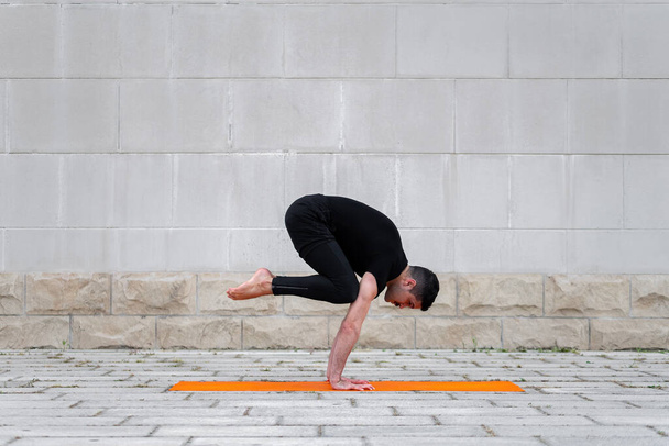 Homem latino praticando ioga ao ar livre em uma cidade, de pé em pose de guindaste no tapete laranja, com parede cinza no fundo
. - Foto, Imagem