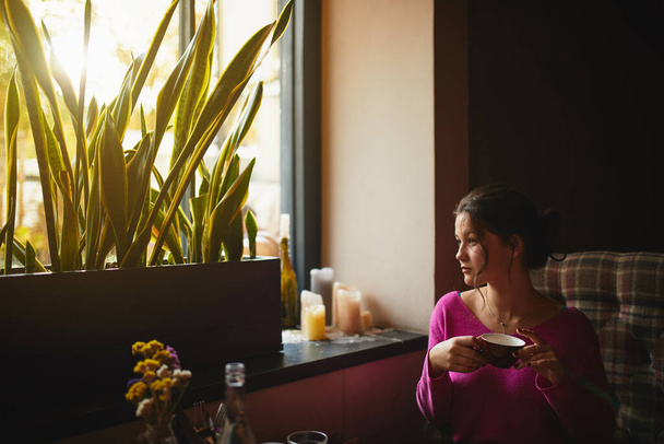 Bonita joven sosteniendo taza roja y sentada en la cafetería mientras sueña
 - Foto, imagen