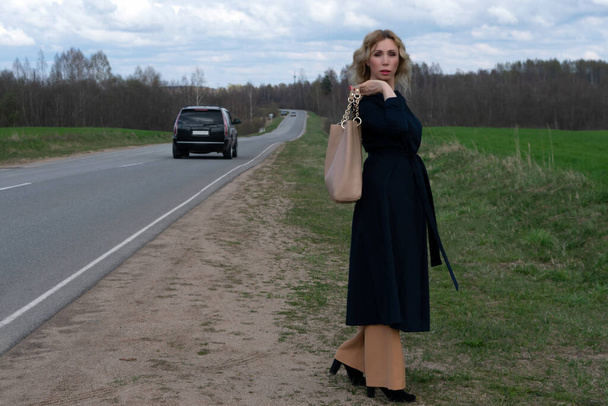 Uma mulher caucasiana de meia-idade em um elegante casaco preto de verão fica sozinha perto de uma estrada no campo contra um céu nublado em um dia de primavera. Copiar espaço para o seu texto ou design
. - Foto, Imagem