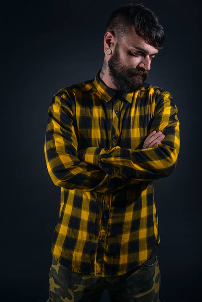 Attrayant homme barbu avec des tatouages pose avec les bras croisés vêtus de chemise à carreaux jaune et noir sur un fond noir - Photo, image