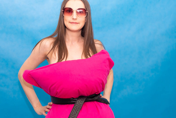 Nette Frau in rosa Kissenkleid und Sonnenbrille auf blauem Hintergrund für verrückte Pillow Challenge, weil sie zu Hause bleiben muss. Caronavirus-Quarantäne. Fashion Girl. Kissenruf wegen Hausdämmung. - Foto, Bild