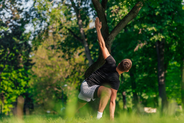 Ελκυστικός καυκάσιος νεαρός άνδρας που τεντώνεται και κάνει ασκήσεις γιόγκα στο πράσινο γρασίδι στο πάρκο - Φωτογραφία, εικόνα