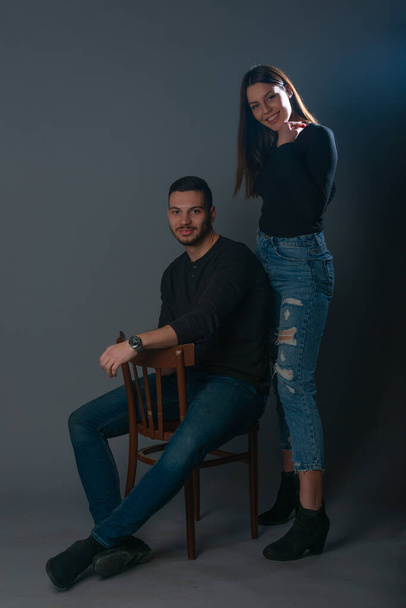 Handsome man sitting on a chair and beautiful woman standing next to him on dark background in a studio - Photo, image