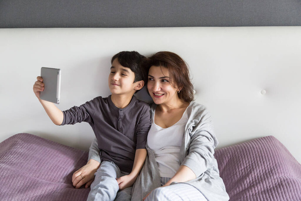 Mother and son having happy family time at home watching movie on tablet, taking a selfie, enjoying domestic life together. Staying home happily, cozy family time concept - Photo, Image