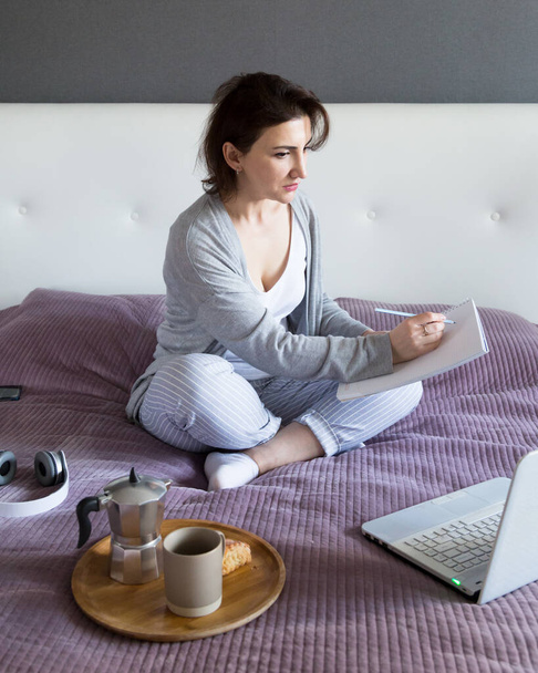 Молода жінка в сірому кольорі працює вдома у своїй спальні, позаштатна робота з ноутбуком і блокнотом. Дистанційне навчання або концепція домашнього офісу
 - Фото, зображення