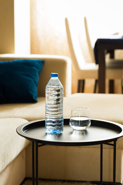 リビングルームのコーヒーテーブルの上に純粋な水と青のプラスチックボトルのガラス。毎日の水消費量、居心地の良い家庭の雰囲気 - 写真・画像