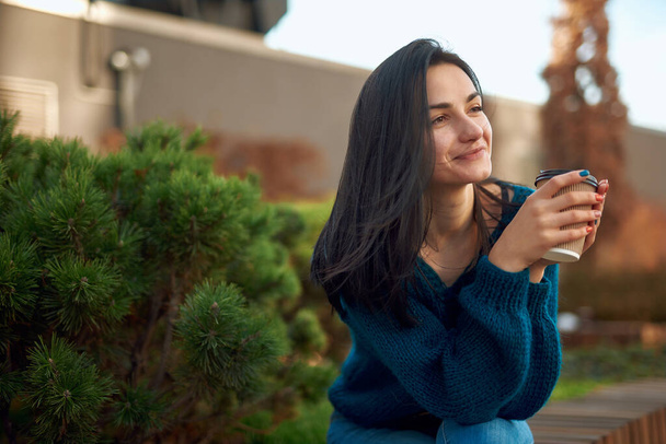 Romantic young lady with lovely smile and far away look holding cup of coffee while sitting on bench in square - Foto, imagen