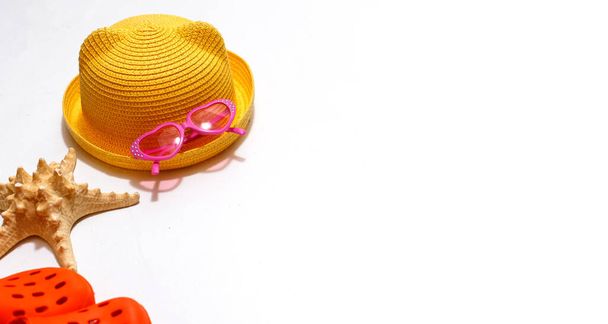 Beach accessories for sun protection: children's yellow straw hat, orange rubber sandals, pink heart-shaped glasses on a white background. Layout with space for text. Ready for travel and beach holidays at sea - Fotó, kép