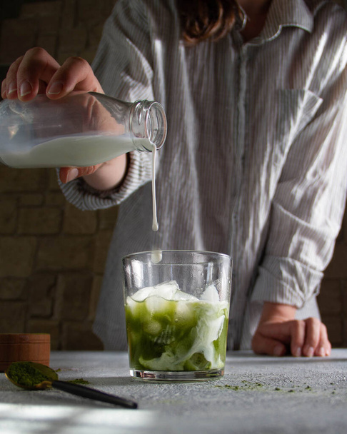 Girl pours milk into matcha green tea in a transparent glass. - Foto, imagen