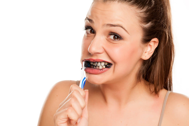 young woman brushing her teeth with black toothpaste of activated charcoal on white background - Fotó, kép
