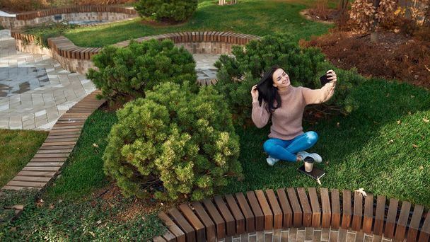 Charming young woman doing photo of herself on a cellphone while sitting cross-legged on grass and touching her long dark hair - Foto, Imagen