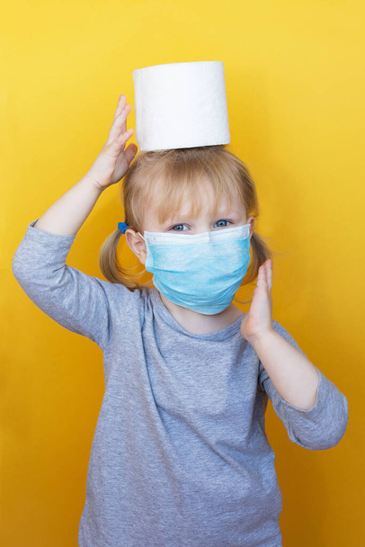 Cute little girl wearing a protective mask holding toilet paper roll on her head. Funny, positive photo. Corona virus scare. Epidemic Precautions. The child is in quarantine. - Fotografie, Obrázek