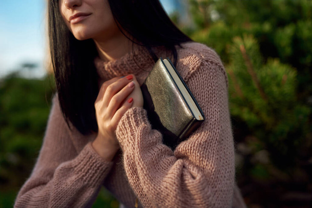 Обрезанный снимок молодой женщины в трикотажном свитере, сжимающей толстый черный дневник, привязанный кожей к ее бресту обеими руками
 - Фото, изображение