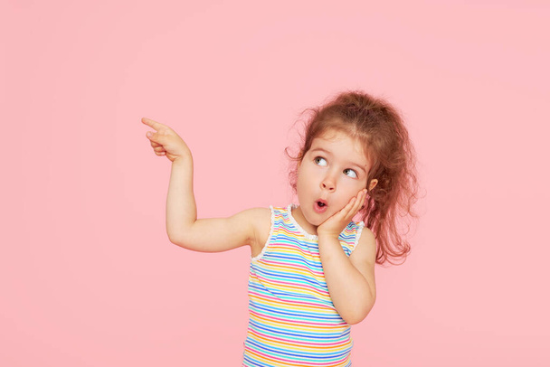 Portrait of surprised cute little toddler girl child over pink background. Looking at camera. Points hands to the left side. Advertising childrens products - Fotografie, Obrázek