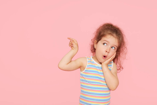 Portrait of surprised cute little toddler girl child over pink background. Looking at camera. Points hands to the left side. Advertising childrens products - 写真・画像