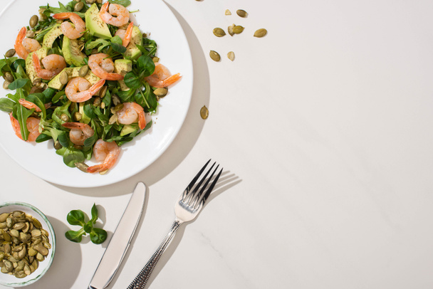 top view of fresh green salad with pumpkin seeds, shrimps and avocado on plate near cutlery on white background - Foto, afbeelding