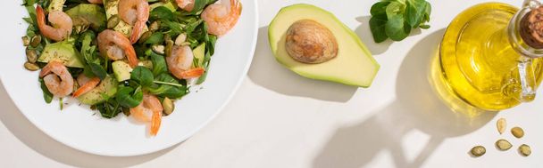 top view of fresh green salad with pumpkin seeds, shrimps and avocado on plate near ingredients on white background, horizontal image - Photo, Image