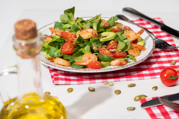 selektiver Schwerpunkt von frischem grünem Salat mit Garnelen und Avocado auf Teller neben Besteck auf karierter Serviette und einem Glas Öl auf weißem Hintergrund - Foto, Bild