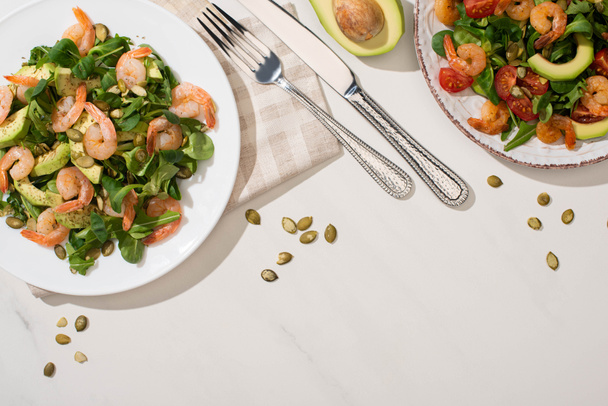 top view of fresh green salad with shrimps and avocado on plates near cutlery on white background - Fotoğraf, Görsel