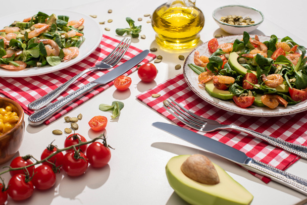 verse groene salade met garnalen en avocado op borden bij het bestek op geruite servetten en ingrediënten op witte tafel - Foto, afbeelding