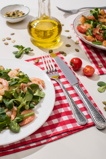 ensalada verde fresca con camarones y aguacate en el plato cerca de cubiertos en servilleta a cuadros e ingredientes sobre fondo blanco
 - Foto, imagen