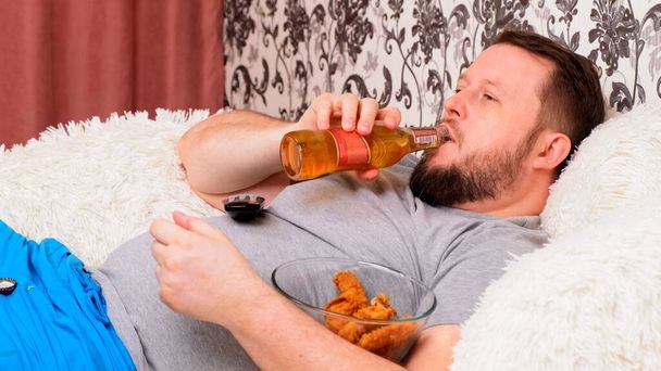 Şişman adam bira içiyor ve sağlıksız yemek yiyor tavuk kanadı yiyor, kanepede televizyon karşısında sıkılıyor. Yetersiz beslenme, evde karantina, alkolizm kavramı.. - Fotoğraf, Görsel
