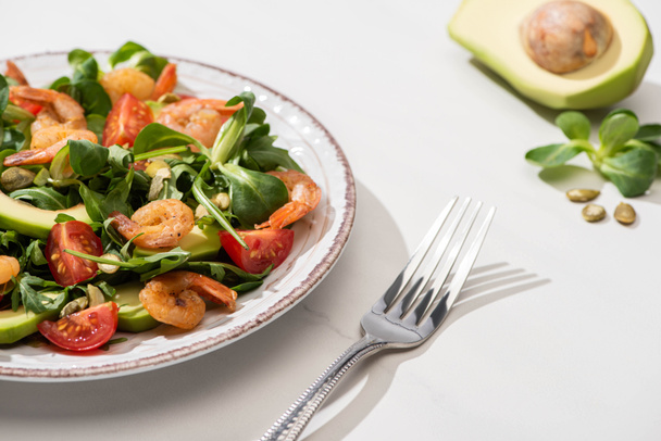 selective focus of fresh green salad with shrimps and avocado on plate near fork and ingredients on white background - Photo, Image