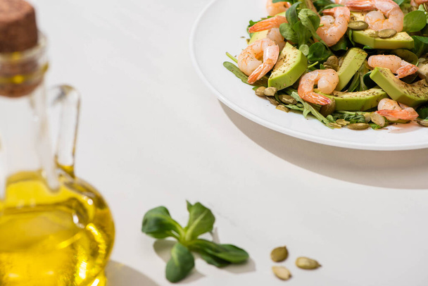 selektywne skupienie świeżej sałatki zielonej z krewetkami i awokado na talerzu w pobliżu oliwy z oliwek na białym tle - Zdjęcie, obraz
